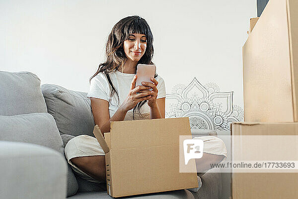 Lächelnde Frau  die Textnachrichten über ihr Smartphone sendet und zu Hause mit einem Karton auf dem Sofa sitzt