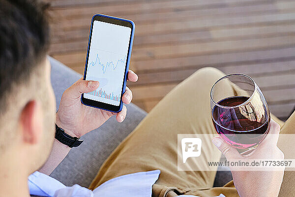 Mann hält Weinglas und Smartphone mit Börsendaten