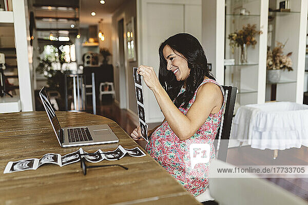 Happy pregnant woman showing ultrasound scans through video call on laptop at home