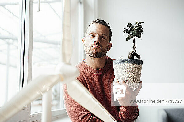 Man holding pot of Bonsai plant looking at wind turbine model at home