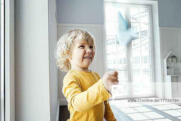 Cute boy playing with origami bird standing at home