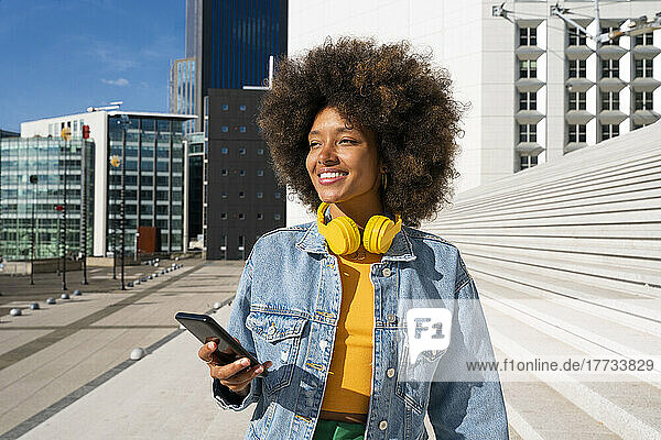 Smiling beautiful young woman wearing wireless headphones standing with mobile phone by steps