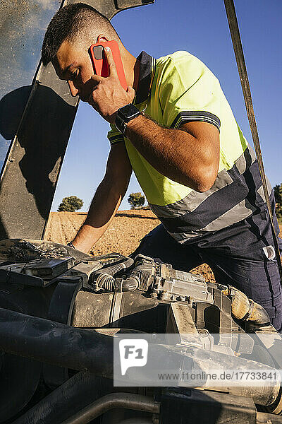 Young farmer talking on mobile phone standing by broken down tractor