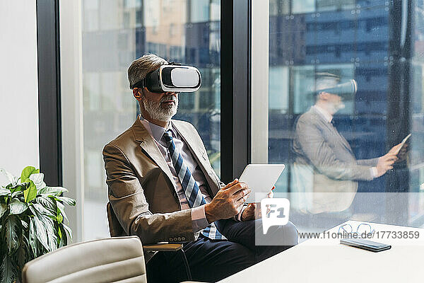 Mature businessman wearing virtual reality simulator sitting with tablet PC at desk