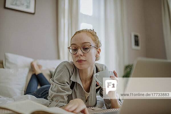Woman holding coffee cup reading book lying on bed at home