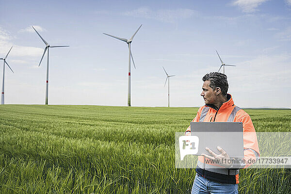 Engineer with laptop standing in front of wind turbines at field