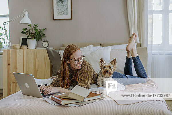 Lächelnde Frau liegt mit Laptop und schaut auf den Hund  der zu Hause im Bett sitzt