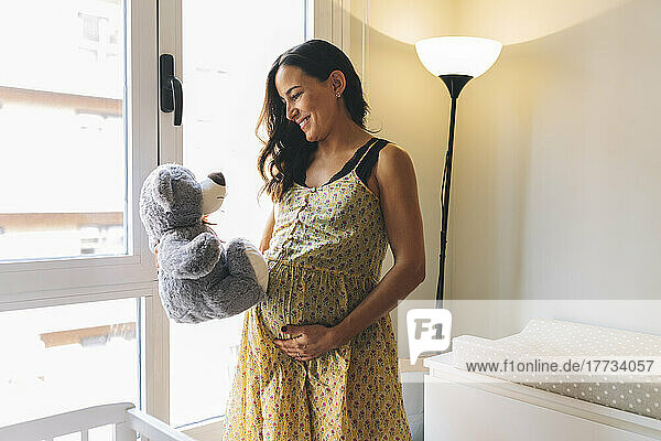 Happy pregnant woman with teddy bear standing by window at home