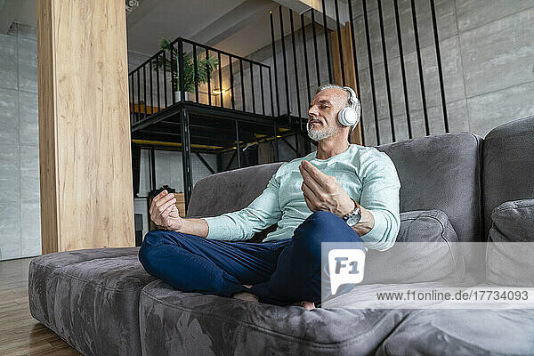 Man listening music through wireless headphones practicing yoga on sofa at home