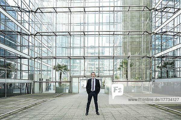 Mature businessman with hands in pockets standing in front of modern glass building