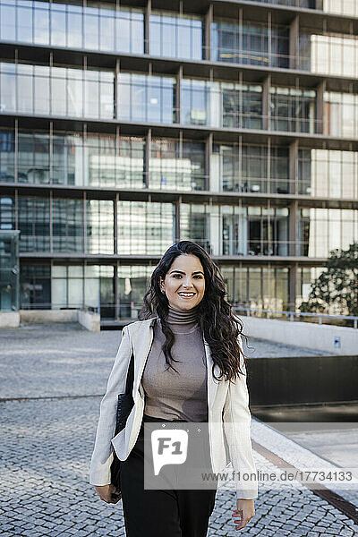 Smiling young businesswoman walking in front of modern office building
