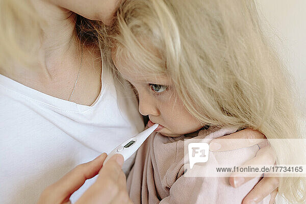 Mother carrying daughter and measuring temperature with thermometer at home