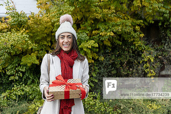 Cheerful young woman wearing knit hat standing with Christmas present at park