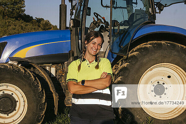 Smiling female farmer standing with arms crossed in front of tractor