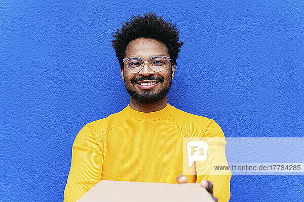 Smiling delivery man giving box in front of blue wall
