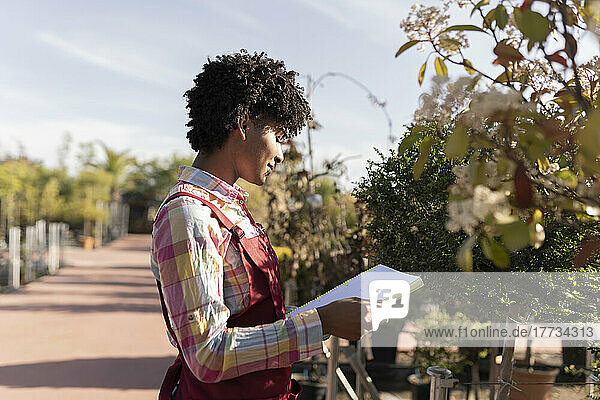 Young gardener holding book standing near plants in nursery