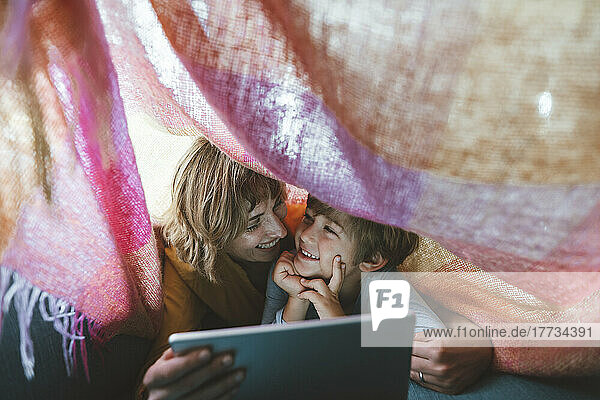 Cheerful mother and son with tablet PC lying under blanket