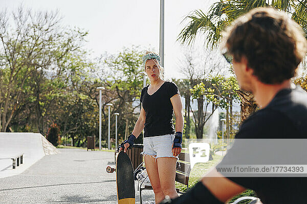 Frau hält Skateboard und blickt Freund an sonnigem Tag an