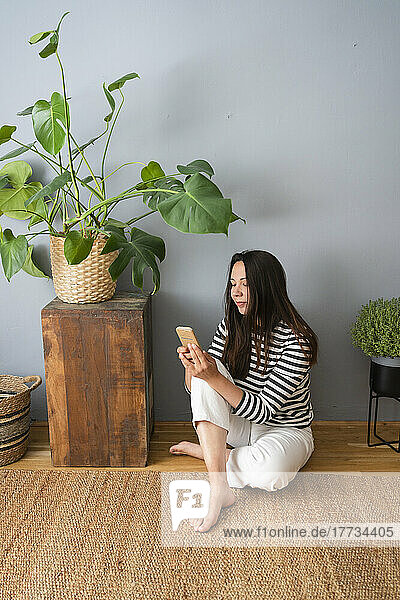 Woman using smart phone sitting on floor in front of gray wall at home