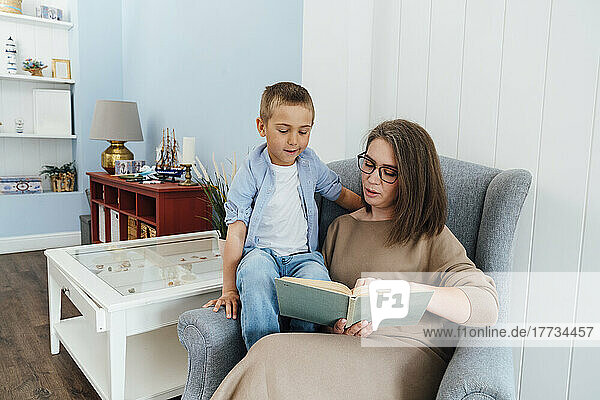 Mother reading book to son sitting on armchair at home
