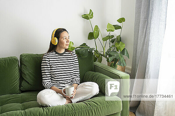 Frau mit Kaffeetasse hört Musik über kabellose Kopfhörer auf dem heimischen Sofa