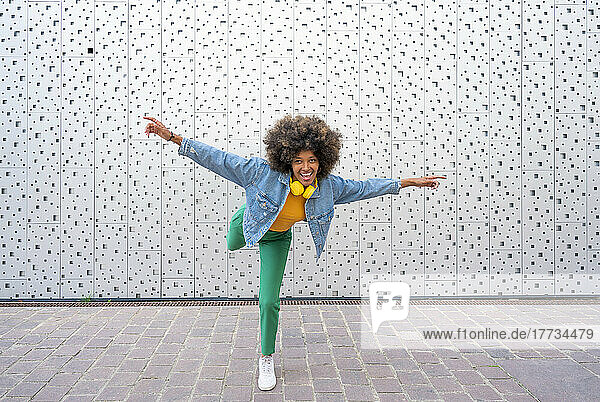 Happy Afro woman with arms outstretched standing on one leg in front of wall