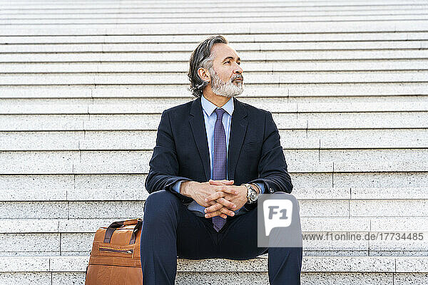 Businessman with hands clasped sitting by bag on steps
