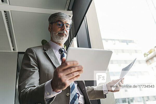 Mature businessman with documents using tablet PC standing by window at office