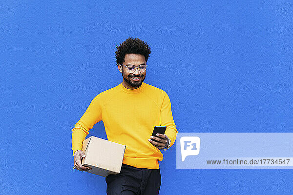 Lächelnder Lieferbote hält Karton mit Smartphone vor blauer Wand