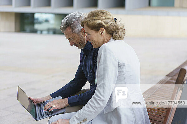 Businessman using laptop sitting by businesswoman on bench