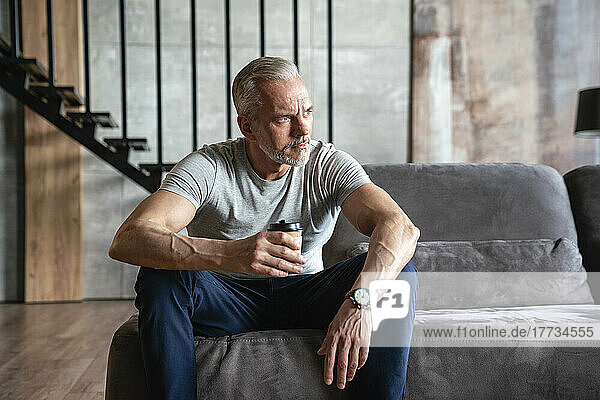 Thoughtful man holding disposable cup sitting on sofa