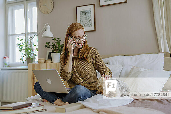 Smiling woman talking on smart phone stroking pet dog in bedroom at home