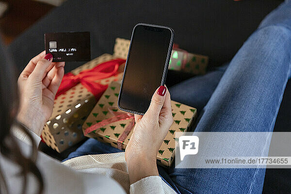 Woman holding credit card doing online shopping through smart phone sitting by Christmas present