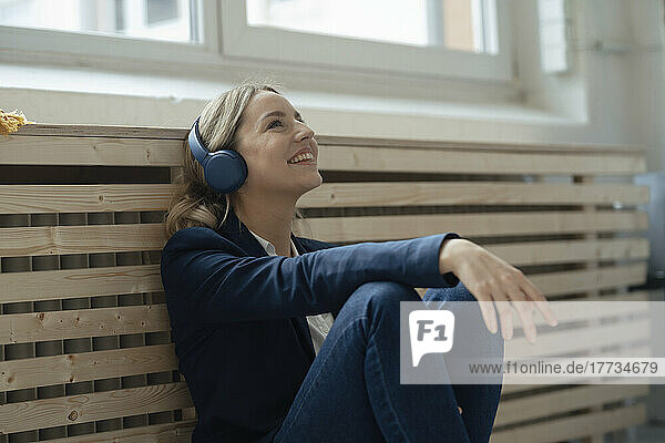 Smiling businesswoman listening music through wireless headphones leaning on radiator in office