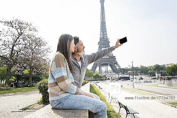 Reifer Mann macht Selfie mit Frau per Smartphone  die an der Wand vor dem Eiffelturm sitzt  Paris  Frankreich