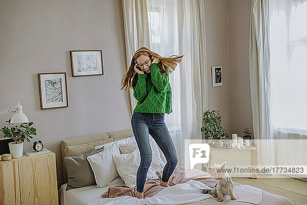 Happy woman listening music through wireless headphones enjoying dance on bed by dog at home