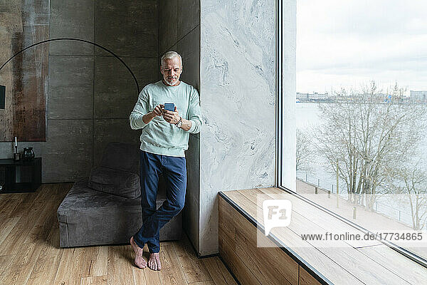Mature man using smart phone standing by window at home