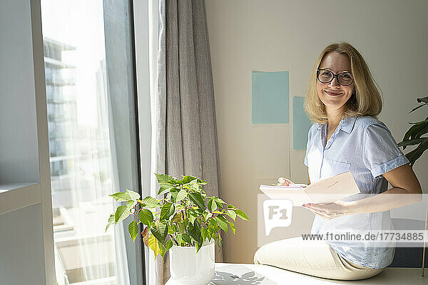 Smiling freelancer wearing eyeglasses sitting with diary on desk in home office
