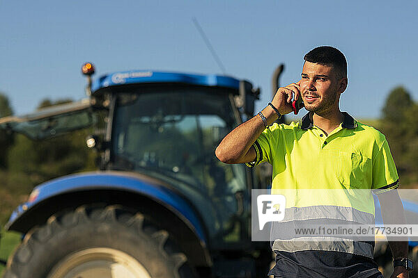 Young farmer talking on smart phone in front of tractor