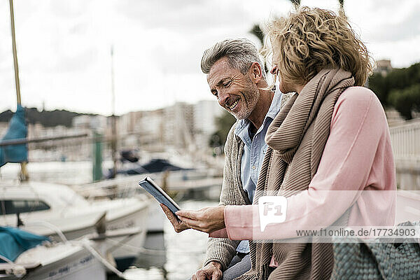 Smiling couple reading book sitting at harbor on vacation