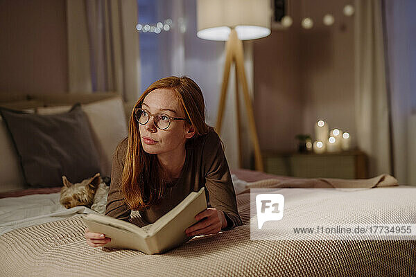 Nachdenkliche Frau mit Brille liegt mit Buch neben Hund zu Hause im Bett
