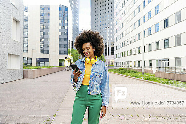 Happy Afro woman wearing wireless headphones using mobile phone standing on footpath