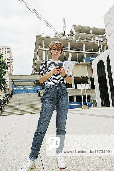 Architect holding blueprints using smart phone standing in front of building