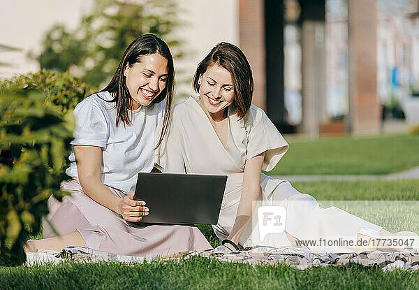 Happy female friends sharing laptop sitting at park on sunny day