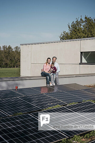 Smiling man and woman with daughter sitting on wall by solar panel