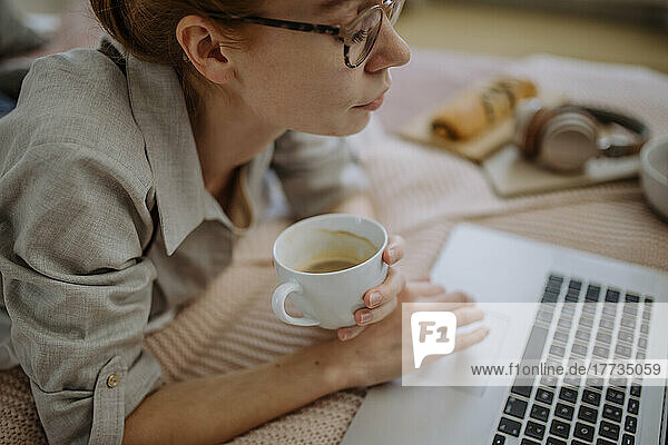 Frau mit Brille und Kaffeetasse hält zu Hause E-Learning über Laptop