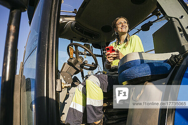 Smiling female farmer holding mobile phone sitting in tractor