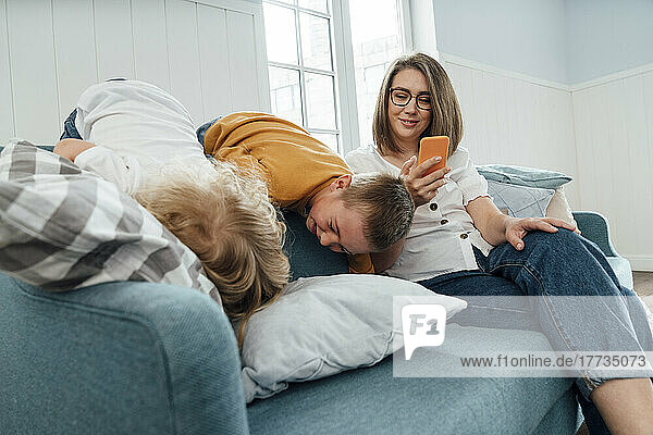Happy woman using smart phone by sons playing on sofa at home