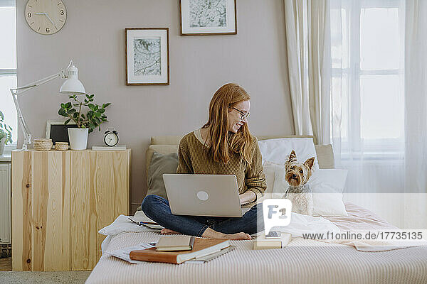 Smiling woman with laptop looking at Yorkshire Terrier sitting on bed at home