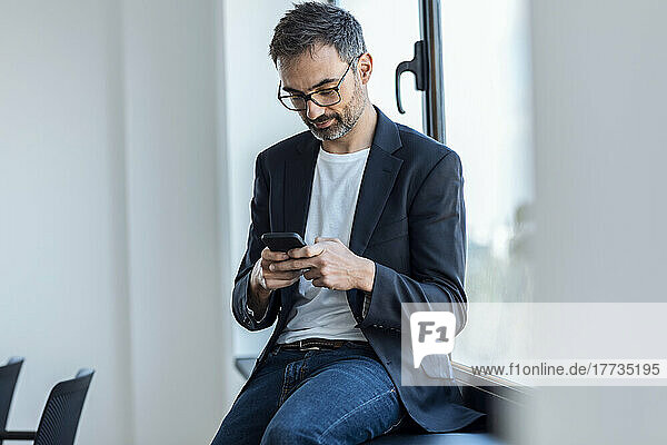 Businessman text messaging through smart phone sitting on window sill in office